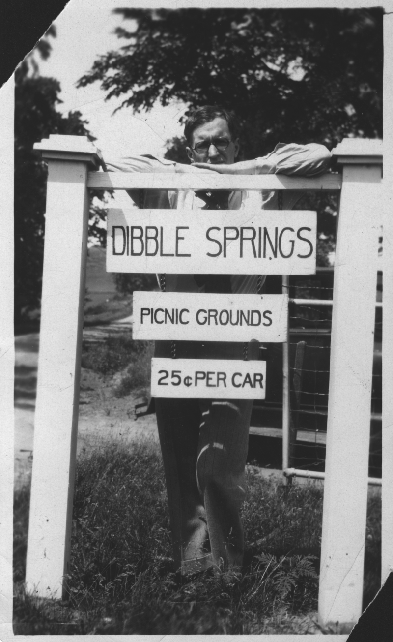 Black and white photo of an unidentified black-haired man standing with his arms crossed on top of a tall white wooden frame from which hangs three signs, in descending order of size. The top sign says 'DIBBLE SPRINGS'; below it a smaller sign says 'Picnic Grounds', and below that an even smaller sign says '25� per Car'.